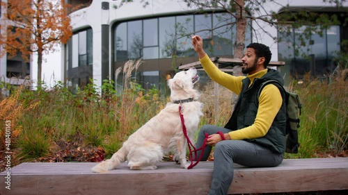 Side view of young man sitting on bench and training his dog outdoors in town.