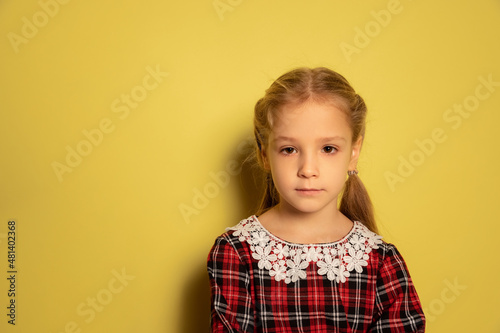 Half-length portrait of cute little girl, pupil wearing plaid dress posing isolated on yellow background. Concept of childhood, emotions, study