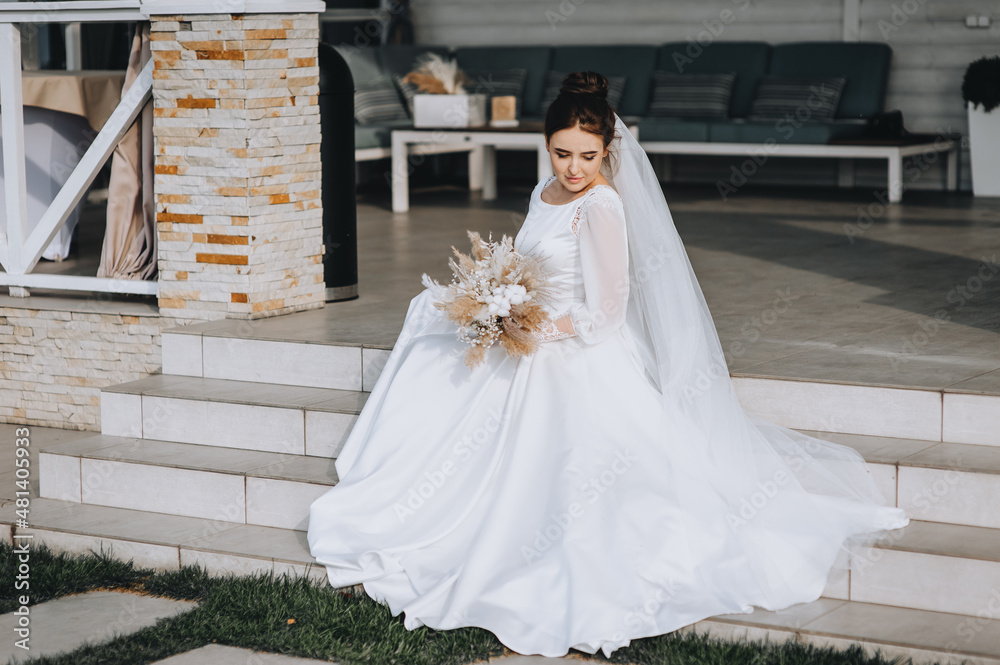 A beautiful, young brunette bride in a white long dress with a bouquet of reeds sits on the steps against the backdrop of a house, a cottage in nature. A beautiful, young brunette bride in a white long dress with a bouquet of reeds sits on the steps against the backdrop of a house, a cottage in nature.