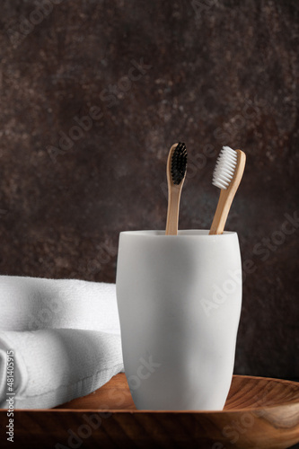 Bamboo toothbrushes in a white glass with dried plant and bath towels on a dark background.