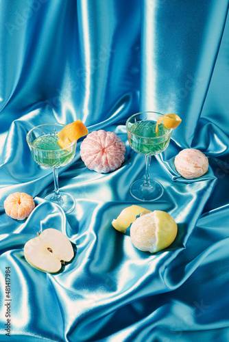 Creative modern still life made against blue satin with exotic drinks in champagne glasses, peeled citrus fruits and apple. Retro minimal background.