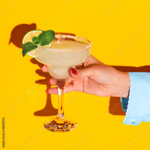 Female hand holding glass with margarita cocktail isolated on bright yellow neon background with shadow. Concept of taste, alcoholic drinks photo