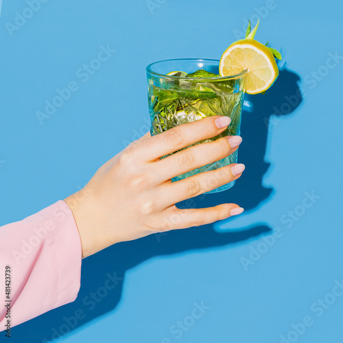 Foto Female hand holding glass with cocktail mojito with lemon isolated on light blue neon background