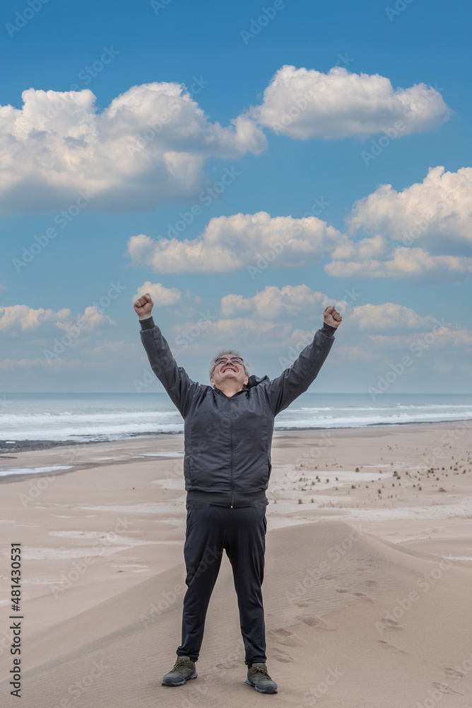 Adult male with gray hair and glasses raising his arms to the sky on top of a dune with the beach and the sea behind