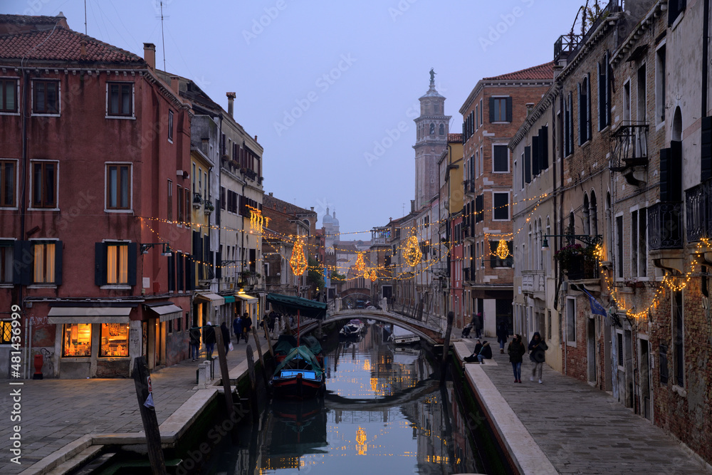 Leute laufen an Wasserkanal in Venedig entlang und weihnachtliche Beleuchtung spiegelt sich im Wasser