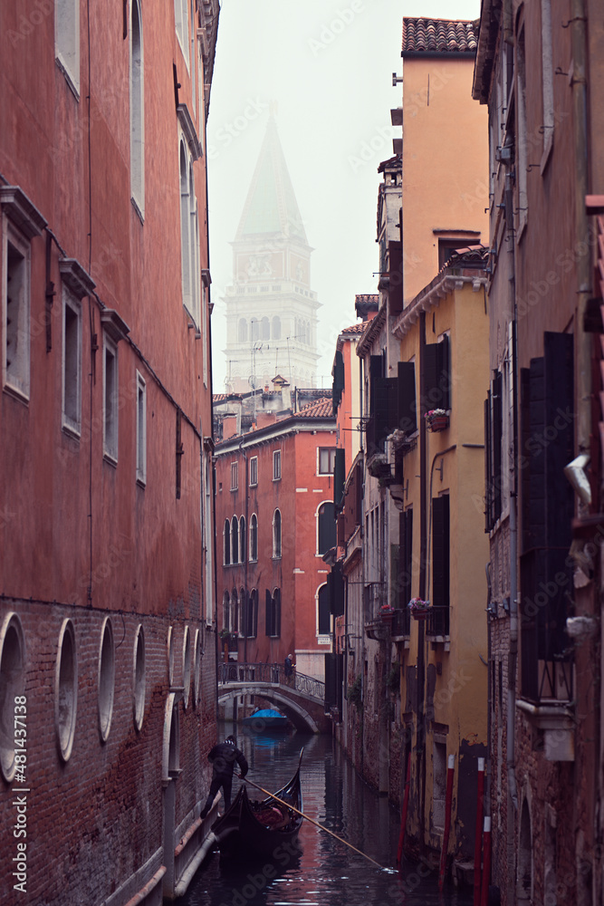 Ein Gondolieri rudert mit seiner Gondel in einem schmalen Kanal zwischen Häusern von Venedig und im Hintergrund ragt der Campanile vom Markusdom über den Dächern hervor