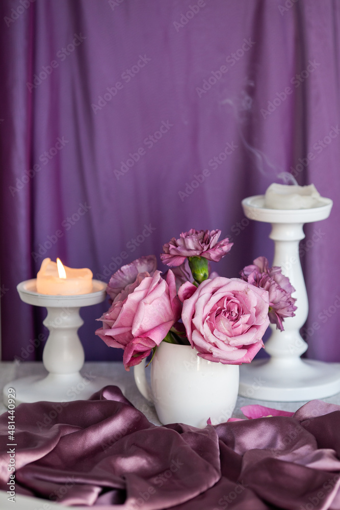 Bouquet of purple roses in a white vase against a background of purple fabric and candles, still life