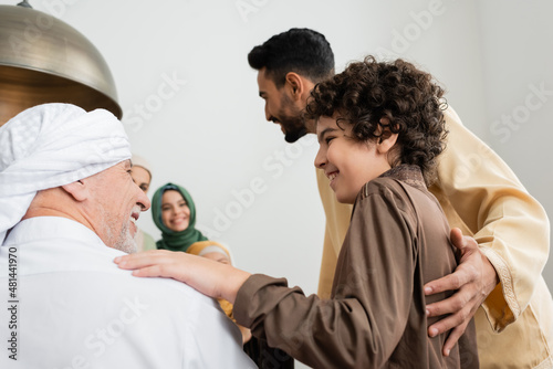 Smiling muslim boy hugging granddad near family at home.
