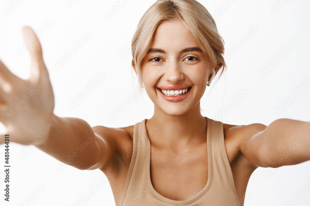 Happy portrait of girl athlete, fitness woman taking selfie in gym clothes, workout of lifestyle blogger, listening music in wireless earphones, standing over white background, smiling Happy portrait of girl athlete, fitness woman taking selfie in gym clothes, workout of lifestyle blogger, listening music in wireless earphones, standing over white background, smiling