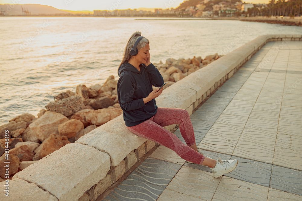 Full-length photo of mature female listening to music and looking on smartphone while sitting on edge of seafront in the evening