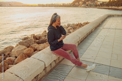 Full-length photo of mature female listening to music and looking on smartphone while sitting on edge of seafront in the evening