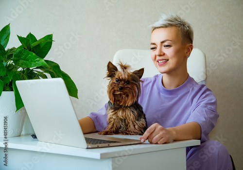 a young woman, blonde with short hair,sits a lady in purple pajamas,near a laptop, and holds a small dog in her arms