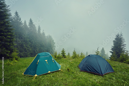 Summer view of small comfortable modern tourist tent on grassy hill under beautiful blue sky on foggy mountains covered with forest background.