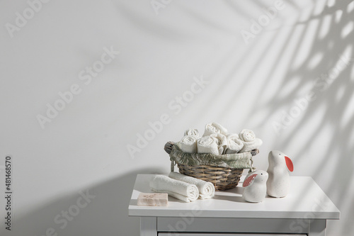 A wicker basket with rolled face towels, soap, two ceramic figurines of hares on a chest of drawers. The shadow of palm leaf. Bathroom decoration for Easter. Place for text photo