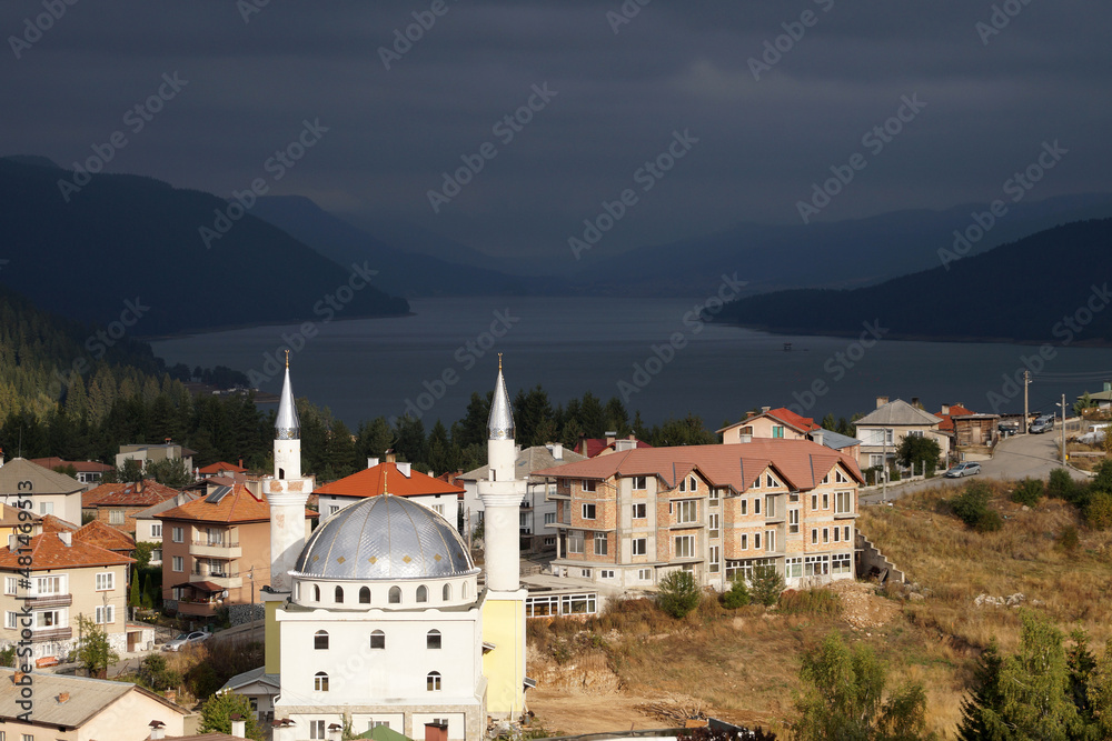 a small town with a mosque in sunlight against a stormy sky, mountains and a lake. Lake Dospat Bulgaria