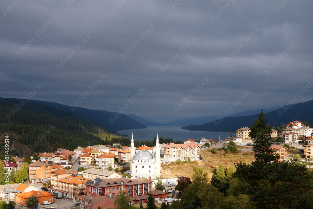 a small town Dospat Bulgaria with a mosque in sunlight against a stormy sky, mountains and a lake