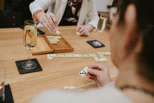 Thin fingers and hands of unrecognisable elderly people possibly women playing dominoes in cozy cafe outdoors lying on table with glasses filled with champagne or wine celebrating their meeting