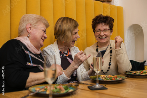 Laughing elderly dark and fair-haired women, having pizza on plates and champagne or wine in glass, dicussing and speaking while celebrating their meeting or holiday in modern cafe with orange walls