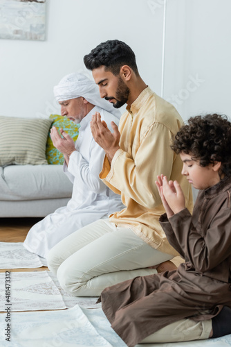 Side view of young muslim man praying near father and son on rugs at home.