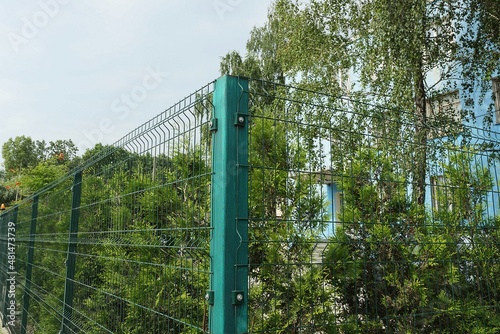 corner of a green wall of a fence made of an iron post and a metal mesh against the background of vegetation and a blue sky