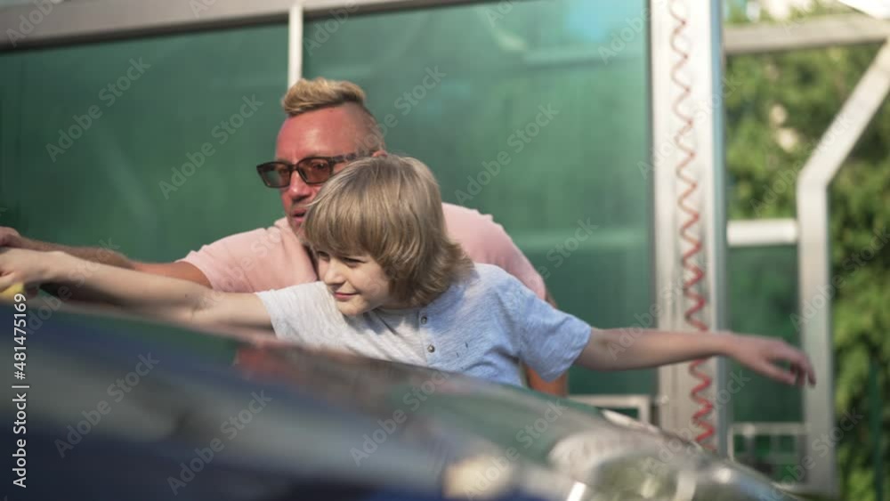 Happy son helping father washing car hood with sponge in slow motion outdoors. Portrait of cute Caucasian boy and handsome man polishing vehicle at car wash service on sunny summer spring day Happy son helping father washing car hood with sponge in slow motion outdoors. Portrait of cute Caucasian boy and handsome man polishing vehicle at car wash service on sunny summer spring day