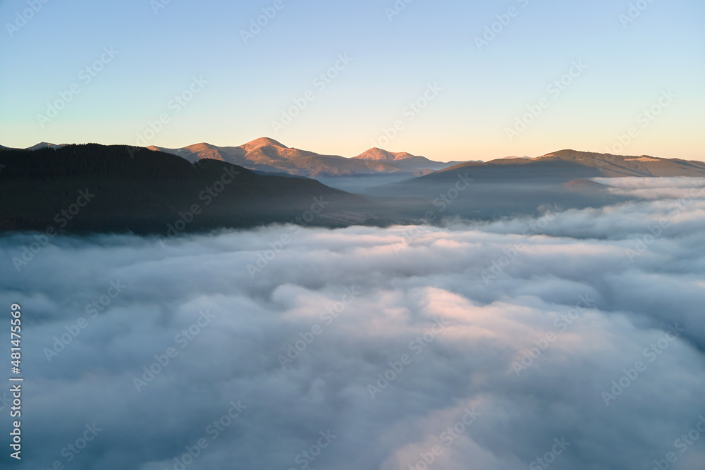 Aerial view of colorful sunrise over white dense fog with distant dark silhouettes of mountain hills on horizon