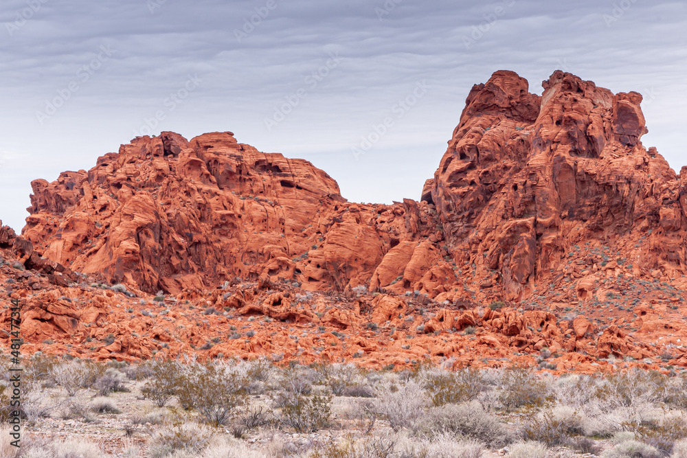 Overton, Nevada, USA - February 24, 2010: Valley of Fire. Red rocky outcrops above beige sand floor with dry bushes under light blue cloudscape.