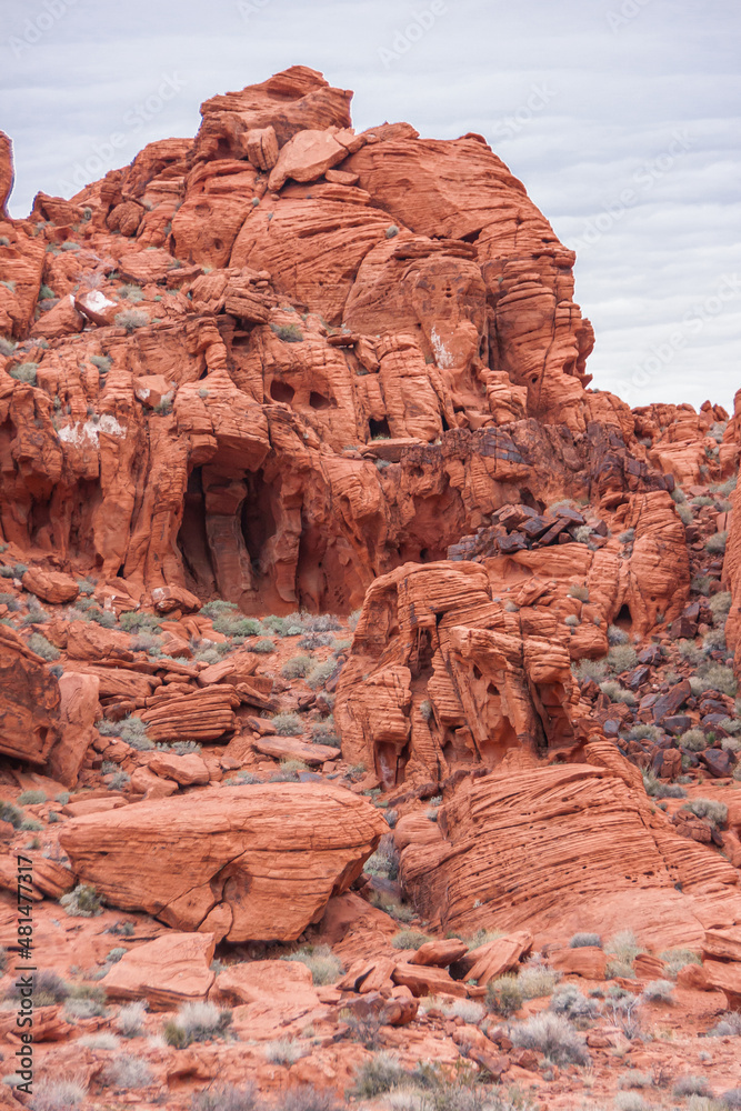 Overton, Nevada, USA - February 24, 2010: Valley of Fire. Closeup portrait of cave-like hole in red rock peak with cracks, black spots and weed crawling up from desert floor. Gray cloudscape. Overton, Nevada, USA - February 24, 2010: Valley of Fire. Closeup portrait of cave-like hole in red rock peak with cracks, black spots and weed crawling up from desert floor. Gray cloudscape.