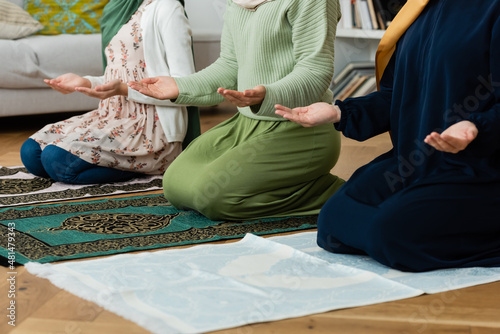 Cropped view of muslim women in traditional clothes praying on rugs at home.