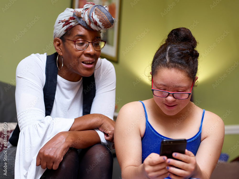 Mother with daughter with Down Syndrome looking at smart phone at home - obrazy, fototapety, plakaty 