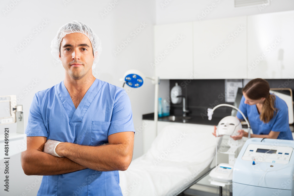 Obraz premium Portrait of positive man doctor of clinic of aesthetic medicine wearing blue overall meeting client