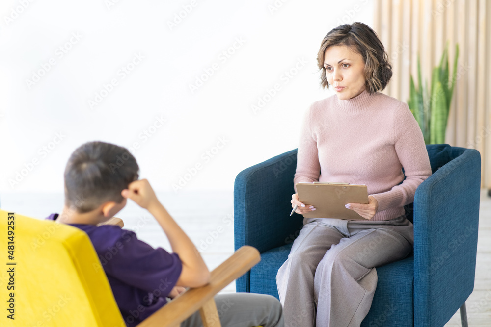 Middle-aged woman psychologist accepts teenager in light cozy office. Writes to his clipboard. Furniture in yellow and blue colors.