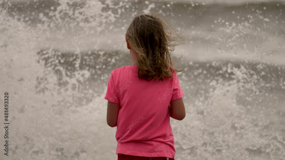 Waves splash up at a little girl as she stands on a beach looking out at the ocean. Waves splash up at a little girl as she stands on a beach looking out at the ocean.