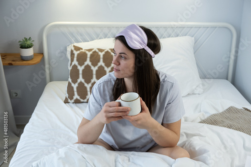 Attractive woman in silk mask having a tea on bed. Portrait of girl enjoying her drink while relaxing at home