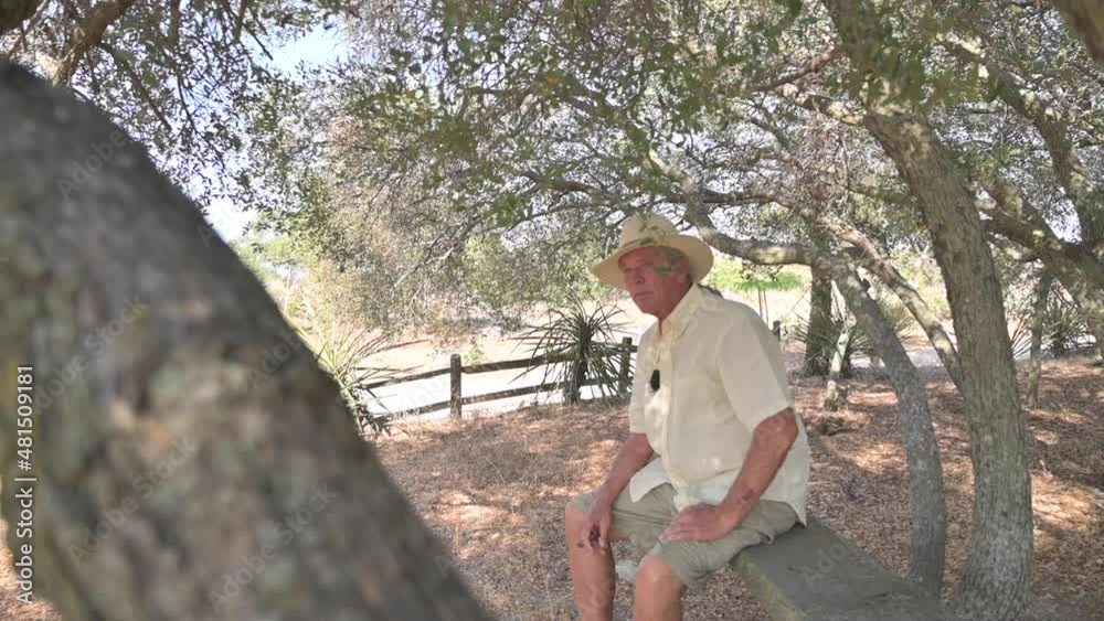 Old man in hat and casual clothing sits on bench under tree, enjoying retirement life. Old man in hat and casual clothing sits on bench under tree, enjoying retirement life.
