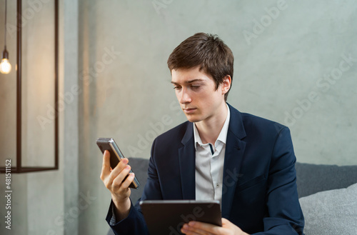caucasian businessman seriously working and looking to tablet and mobile phone, wear suit, sitting, light and space at background. Young man work business with home office background and copy space