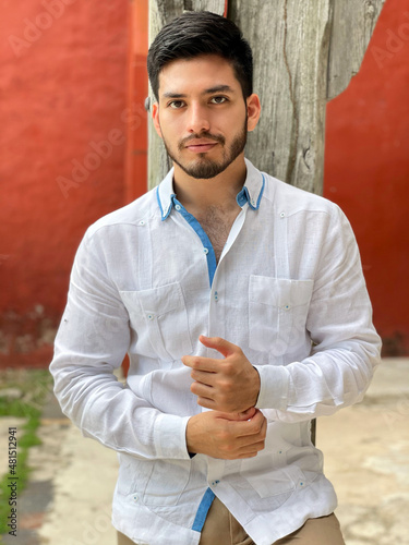 Young model wearing an elegant white guayabera with blue details, traditional linen shirt from southeastern Mexico. Model looking straight ahead with elegant attitude.