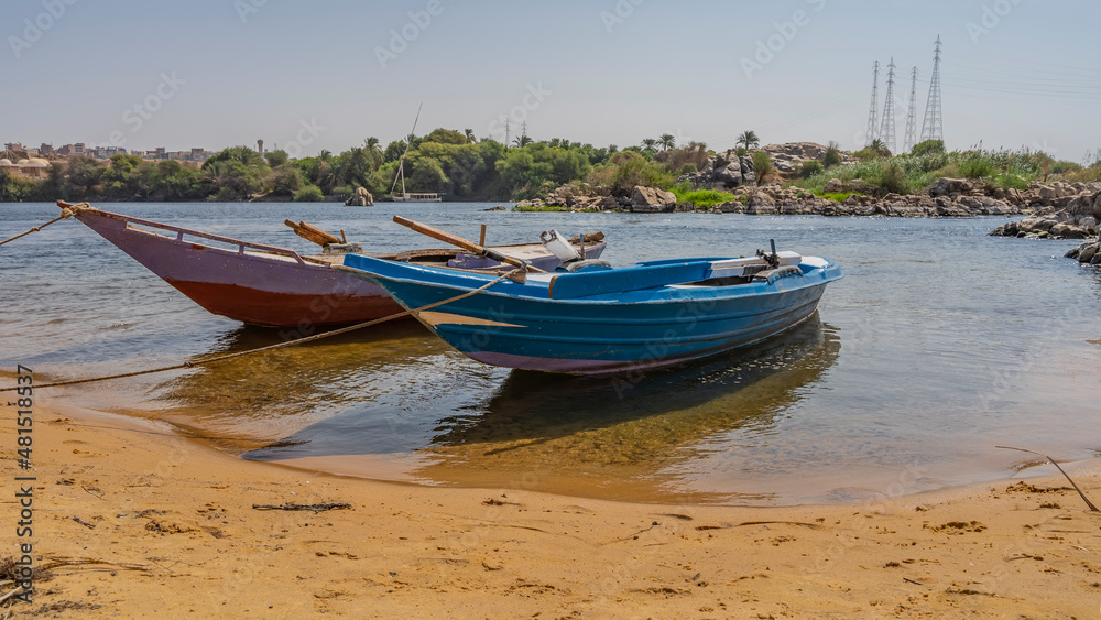 Two bright wooden boats are tied up near the bank of the Nile. There is yellow sand on the beach. Shadows and reflections on clear water. Green vegetation, boulders against the blue sky. Egypt. Aswan