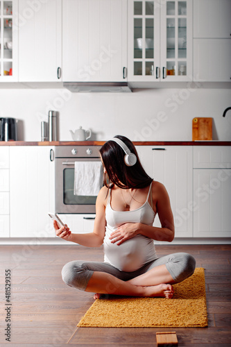 Pregnant doing workout on orange fitness mat. Pragnant yoga at home in the kitchen. Pregnant in white headphones.