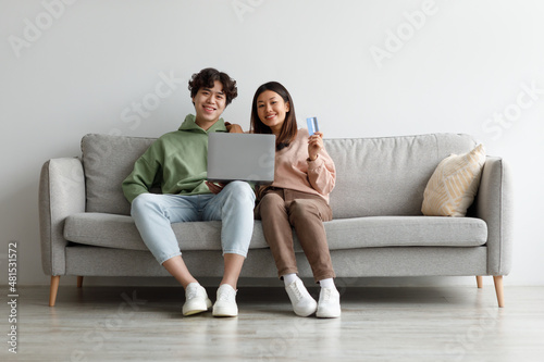 Online shopping. Asian couple sitting on couch at home, using laptop and credit card, purchasing goods in web store