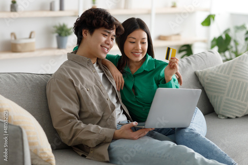 Positive Asian family doing online shopping with credit card and laptop, sitting on sofa and cuddling at home © Prostock-studio