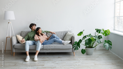 Positive young Asian couple relaxing on sofa, cuddling and looking out window in living room, free space