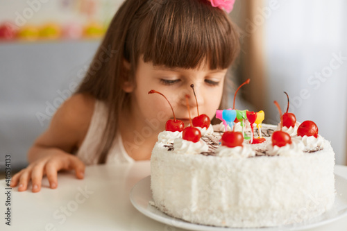 Image of pretty little dark haired girl celebrating birthday smelling her cake  keeps eyes closed  wants to taste and enjoy delicious dessert  happy childhood.
