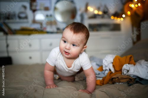 funny european chubby baby crawling among clothes on bed, children's clothes and conscious consumption, lot of clothes and child, big pile of clean clothes and baby playing, childhood and emotions