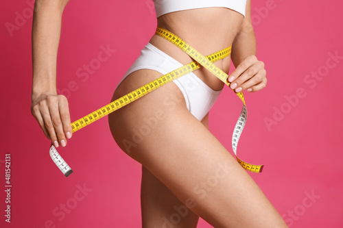 Young woman measuring waist with tape on pink background, closeup