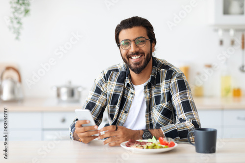 Cheerful middle-eastern man enjoying breakfast, holding smartphone