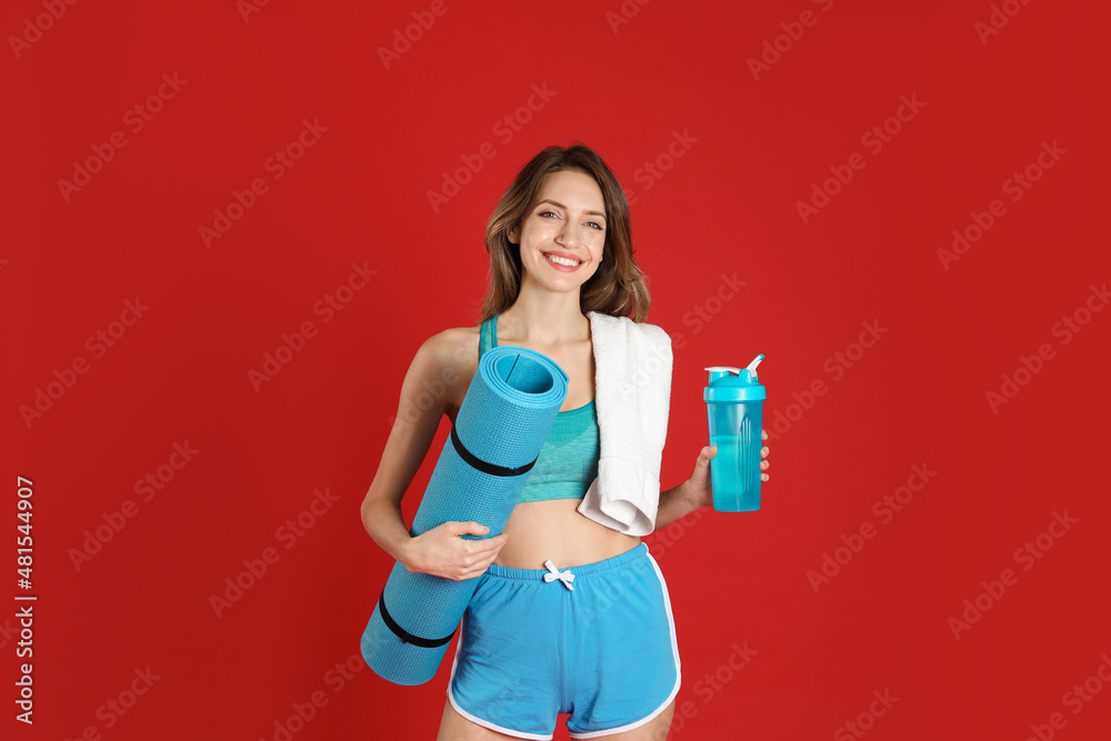 Beautiful woman with yoga mat, towel and shaker on red background