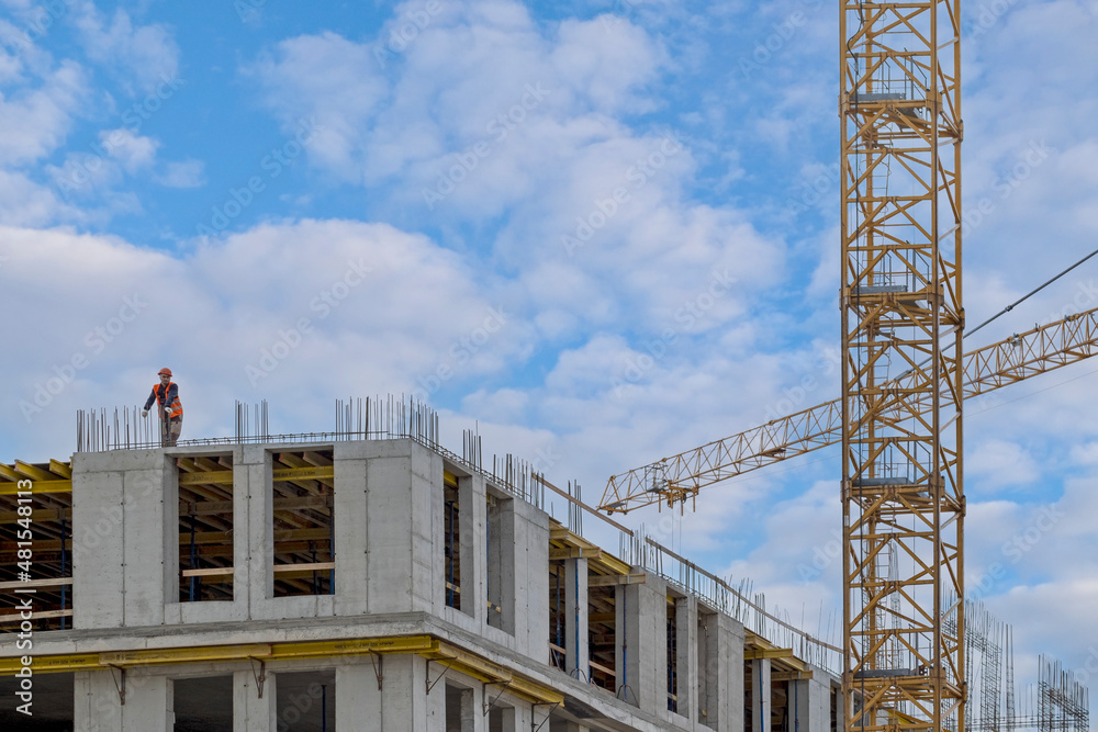 Close-up view of construction of a modern building. A construction worker in a helmet and an orange special vest stands on top and monitors the progress of work.