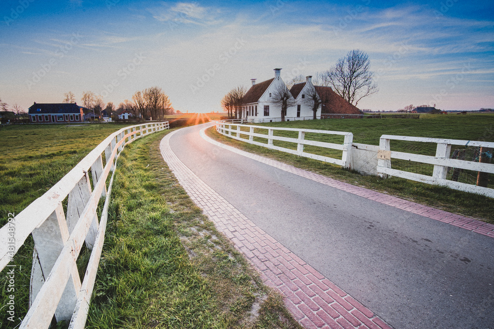 Country road in dutch countryside with white fence leading towards an old farmhouse in Holland during an atmospheric sunset