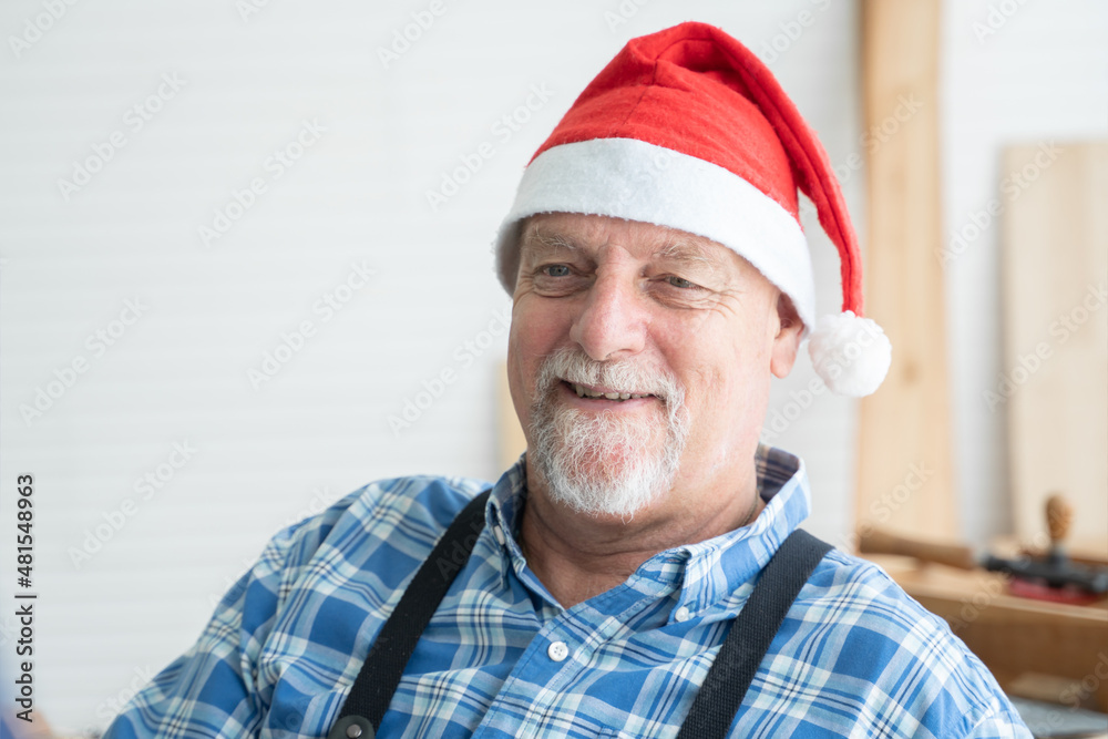 Portrait of happy Caucasian senior carpenter kind man with white beard and mustache wearing santa hat smiling with handmade wooden work on background at workshop on Christmas holiday Portrait of happy Caucasian senior carpenter kind man with white beard and mustache wearing santa hat smiling with handmade wooden work on background at workshop on Christmas holiday